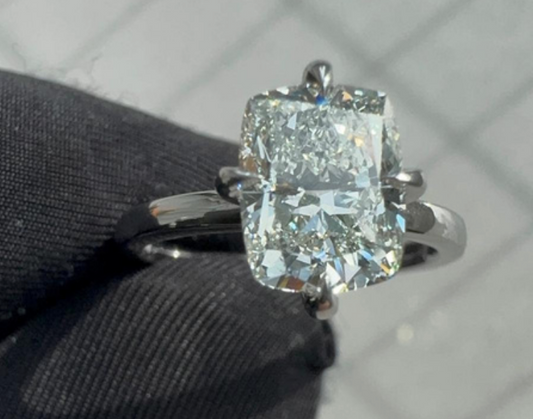 Large cushion-cut diamond solitaire ring held between gloved fingers, featuring a brilliant white stone set in a polished silver or platinum four-prong setting.