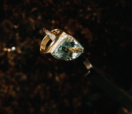 Unique pear-cut aquamarine cocktail ring with a light blue-green stone set in a mixed gold and silver asymmetric mounting, photographed against a moody dark earthy background.