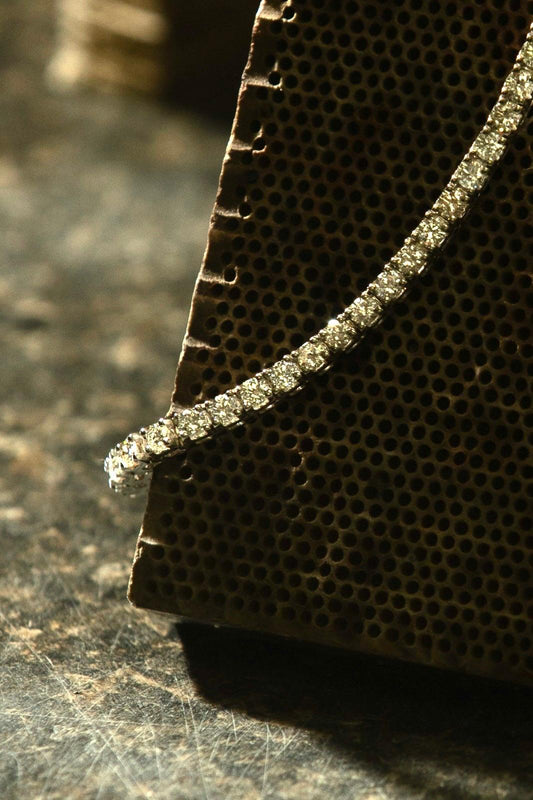 Macro close-up of a diamond tennis bracelet draped over a jeweler's honeycomb soldering block on a scratched metal surface, highlighting the individual stone settings in dramatic side lighting.