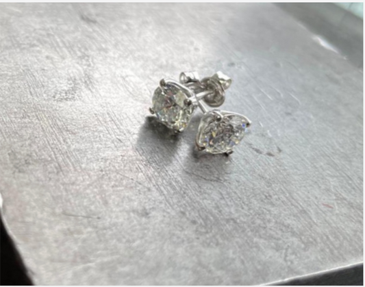 A pair of classic diamond stud earrings with sparkling cushion-cut stones set in silver four-prong settings, photographed on a brushed metal surface.