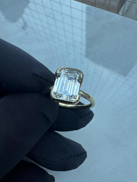Large emerald-cut diamond solitaire ring with a sleek yellow gold bezel setting, held by a gloved hand, showcasing the stone's step-cut clarity and modern minimalist design.