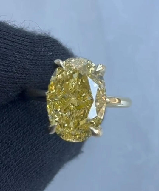 Striking oval-cut fancy yellow diamond solitaire ring held by a gloved hand, set in a yellow gold four-prong setting against a soft blue-grey background, showcasing intense canary yellow brilliance.